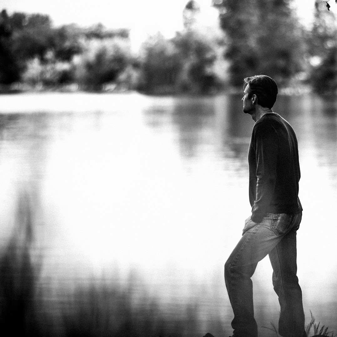 Black and white photo of a man standing and looking at a lake - Mastering Alchemy